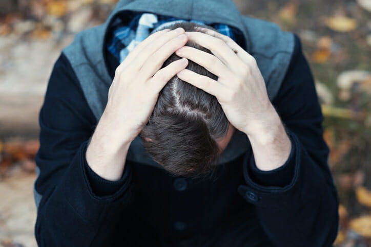 Young man covers his face with his hands in grief or pain. He is sitting outdoor.